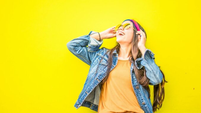 Smiling woman in sunglasses stands against vibrant yellow wall, radiating happiness.